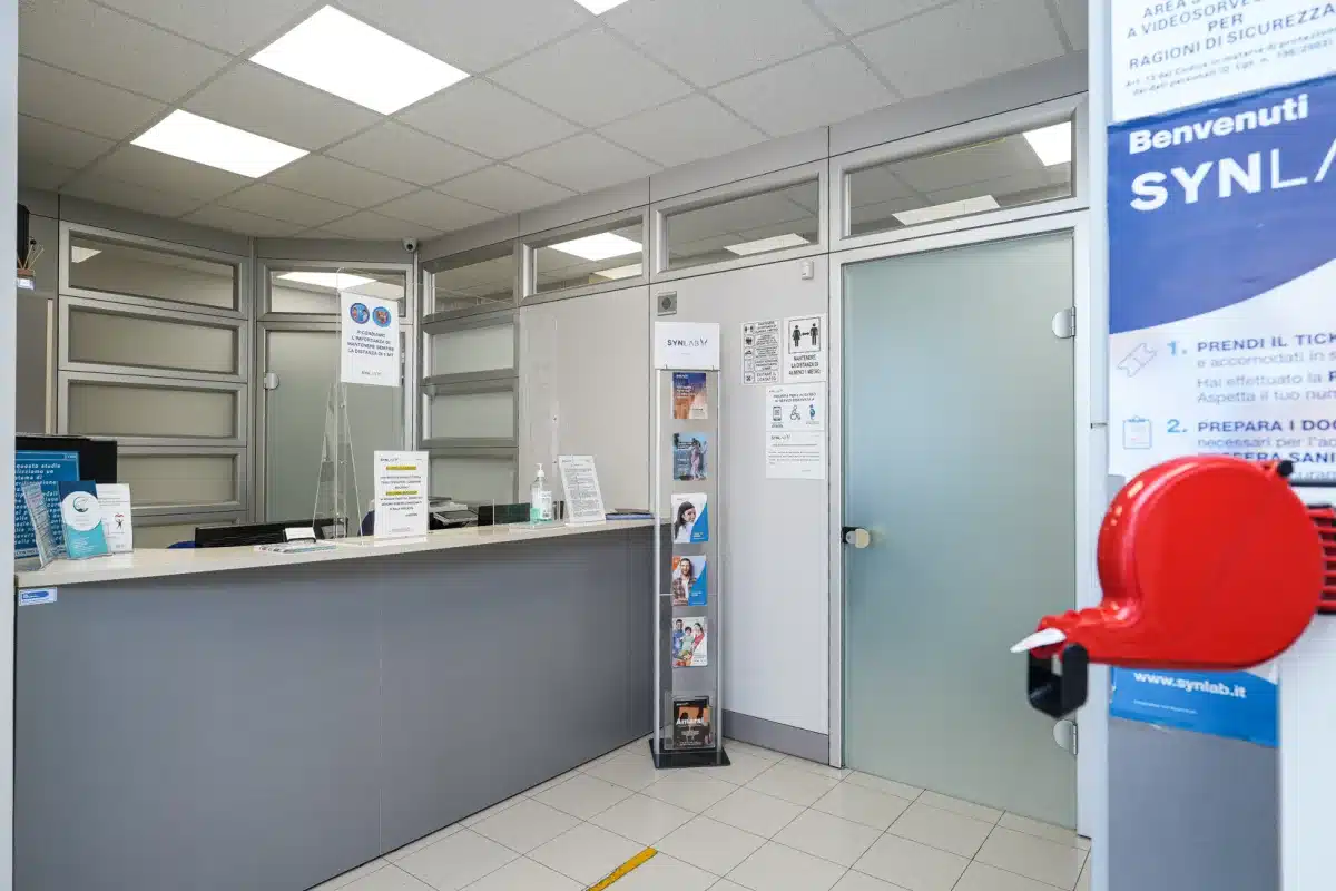 Area di accettazione del punto prelievi Synlab all'interno di Vielle: il bancone della reception, il totem informativo e il cartello di benvenuto Synlab